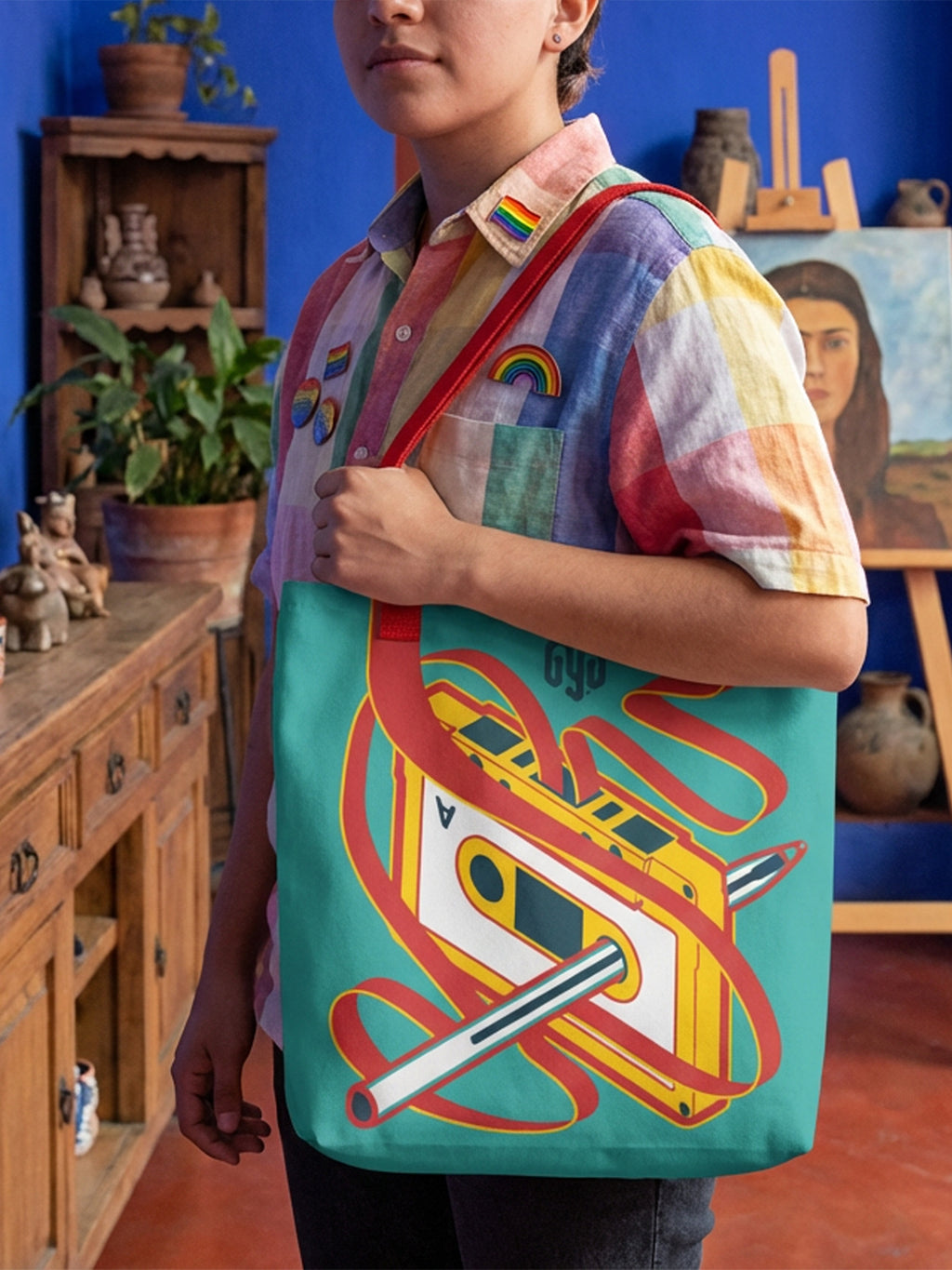 Tote bag en tonos amarillo, rojo y verde, llevada por una persona con estética Pride en una casa de estilo mexicano; diseño de cassette y pluma Bic rebobinando cinta, efecto 3D con asas.