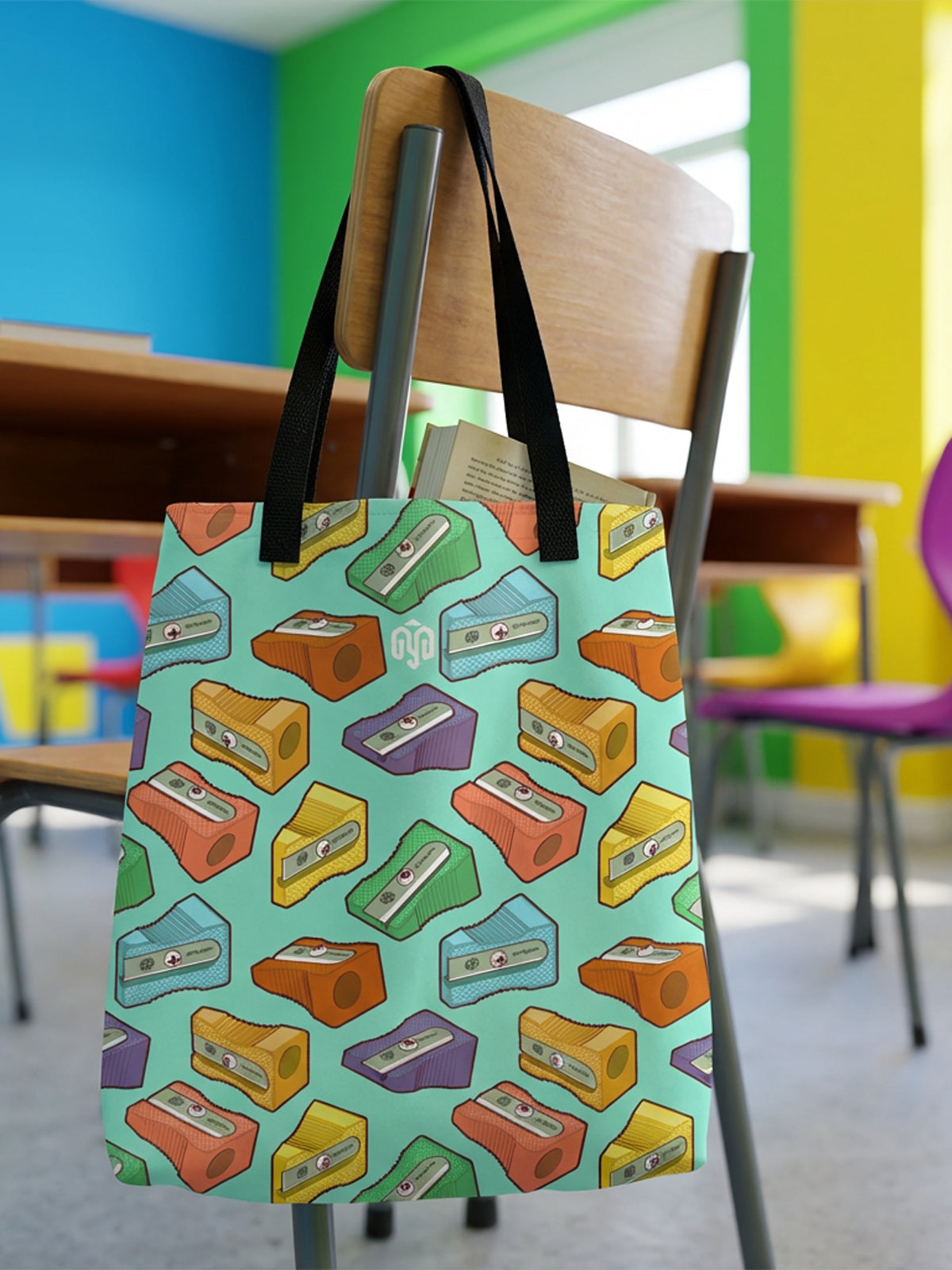 Tote bag con estampado de sacapuntas en tonos pastel, colgada del respaldo de una banca escolar en un salón de clases.